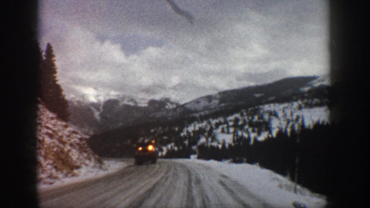 1961 Aspen-area 8mm frame showing a snowy road through mountain forest as a car approaches with headlights on.
