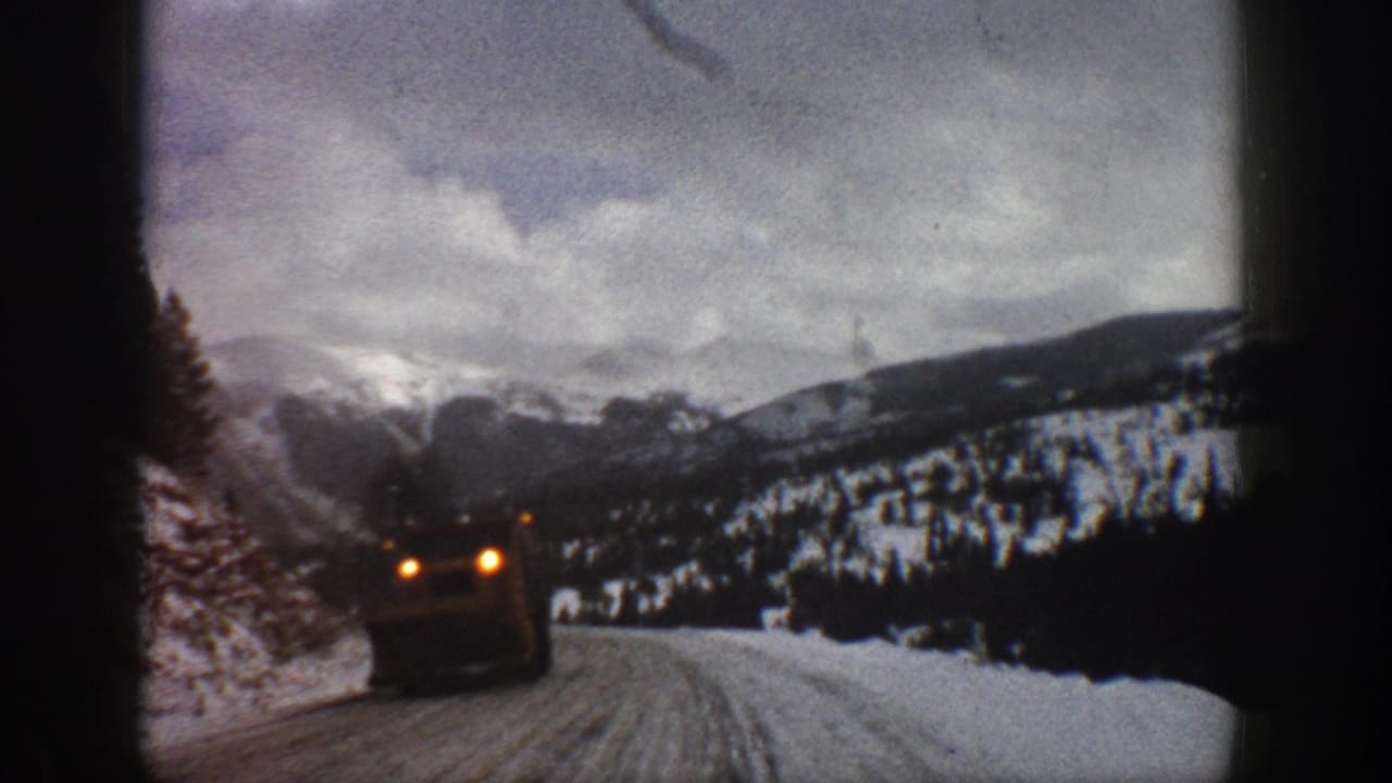 1961 Aspen-area 8mm frame with a car passing close on a snow-packed mountain road beneath cloudy winter peaks.