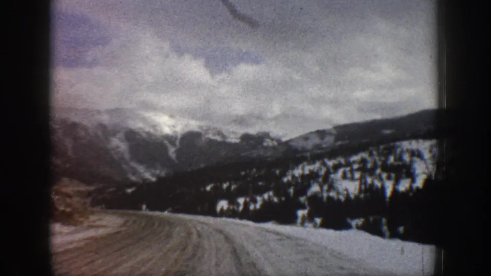 1961 Aspen-area 8mm frame looking down a snow-lined mountain road toward forested slopes and cloudy peaks.