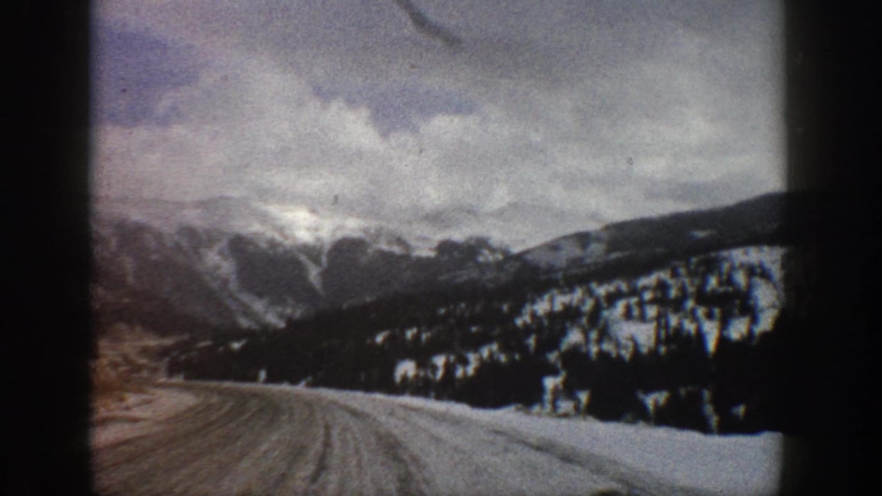 1961 Aspen, Colorado home-movie frame showing an icy road bending through snow-covered mountain terrain and dark evergreen forest.