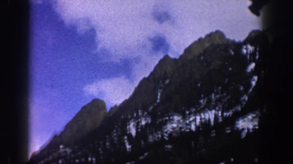 1961 Aspen, Colorado 8mm frame with a rugged snow-dusted mountain ridge and blue-purple color shift in the cloudy sky.