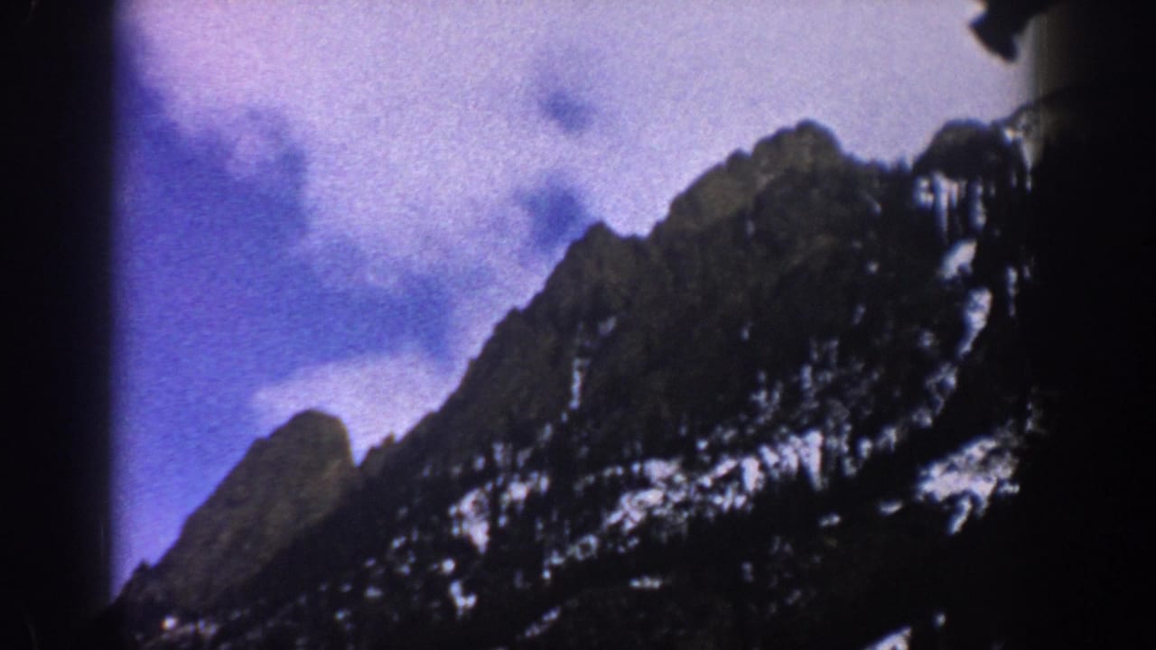 1961 Aspen-area 8mm frame showing rugged snow-patched mountains and a cloudy sky from a moving travel viewpoint.