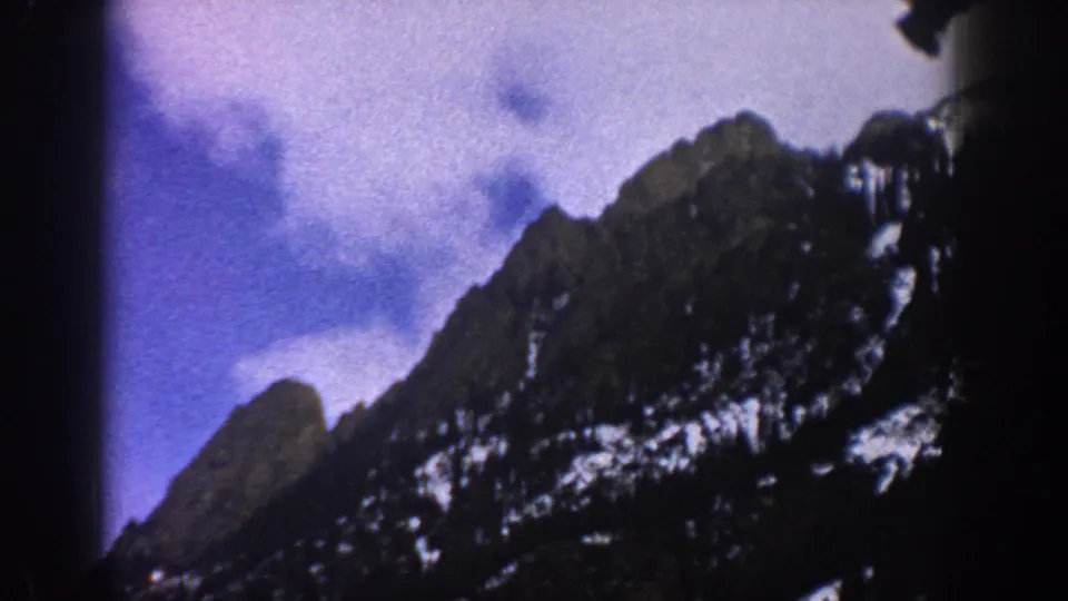 1961 Aspen-area 8mm frame showing rugged snow-patched mountains and a cloudy sky from a moving travel viewpoint.
