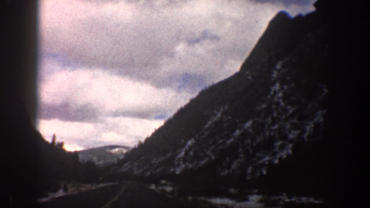 1961 Aspen, Colorado travel footage looks down a road through a narrow mountain valley with snow on the slopes.