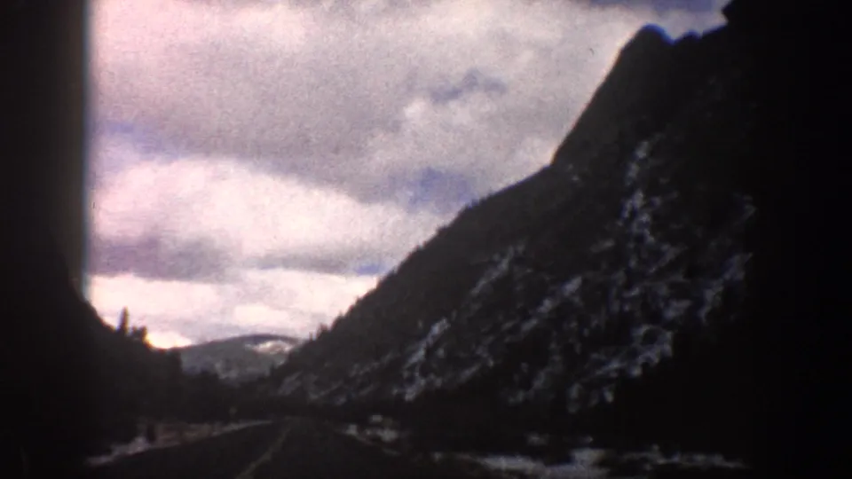 1961 Aspen, Colorado travel footage looks down a road through a narrow mountain valley with snow on the slopes.
