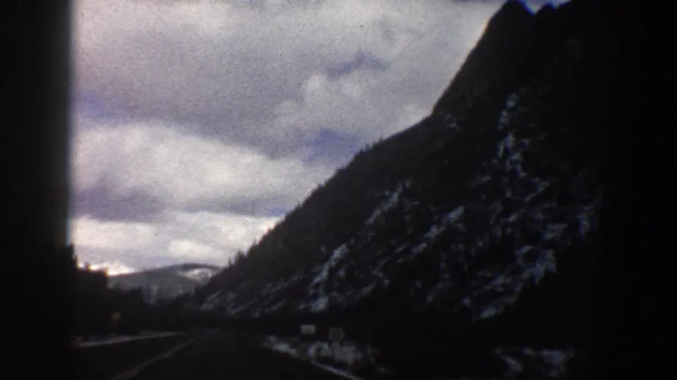 Late frame from the 1961 Aspen-area mountain drive, with the road receding below dark slopes and a cloudy sky.