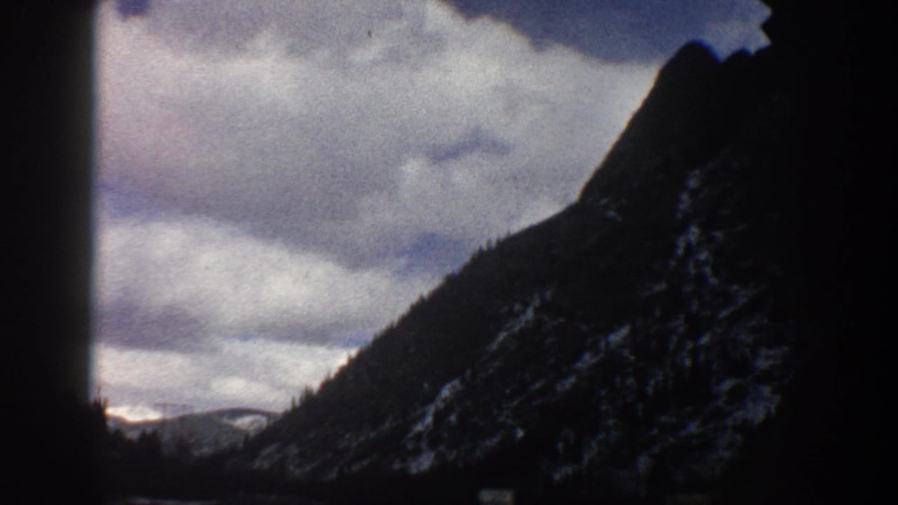 Aspen, Colorado 1961 travel footage frame with a mountain slope, cloud cover, and dark car-window silhouettes.