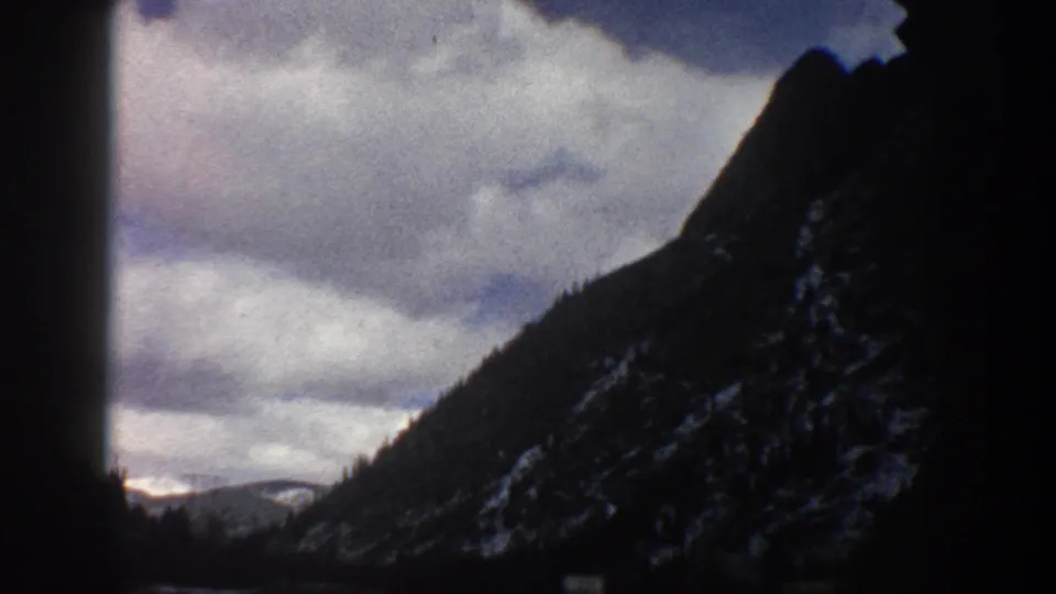 Aspen, Colorado 1961 travel footage frame with a mountain slope, cloud cover, and dark car-window silhouettes.