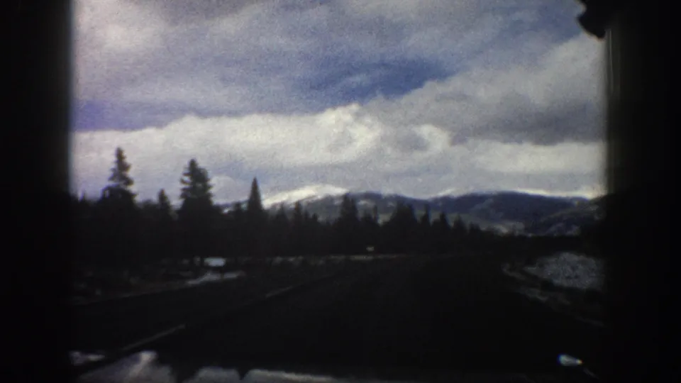 1961 Aspen-area road view with evergreen silhouettes, distant snow-dusted hills, wet roadside ground, and cloudy sky.