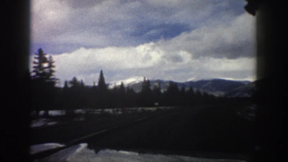 Aspen, Colorado 1961 road-travel frame showing a dark road, tree line, distant snowy hills, and overcast sky.