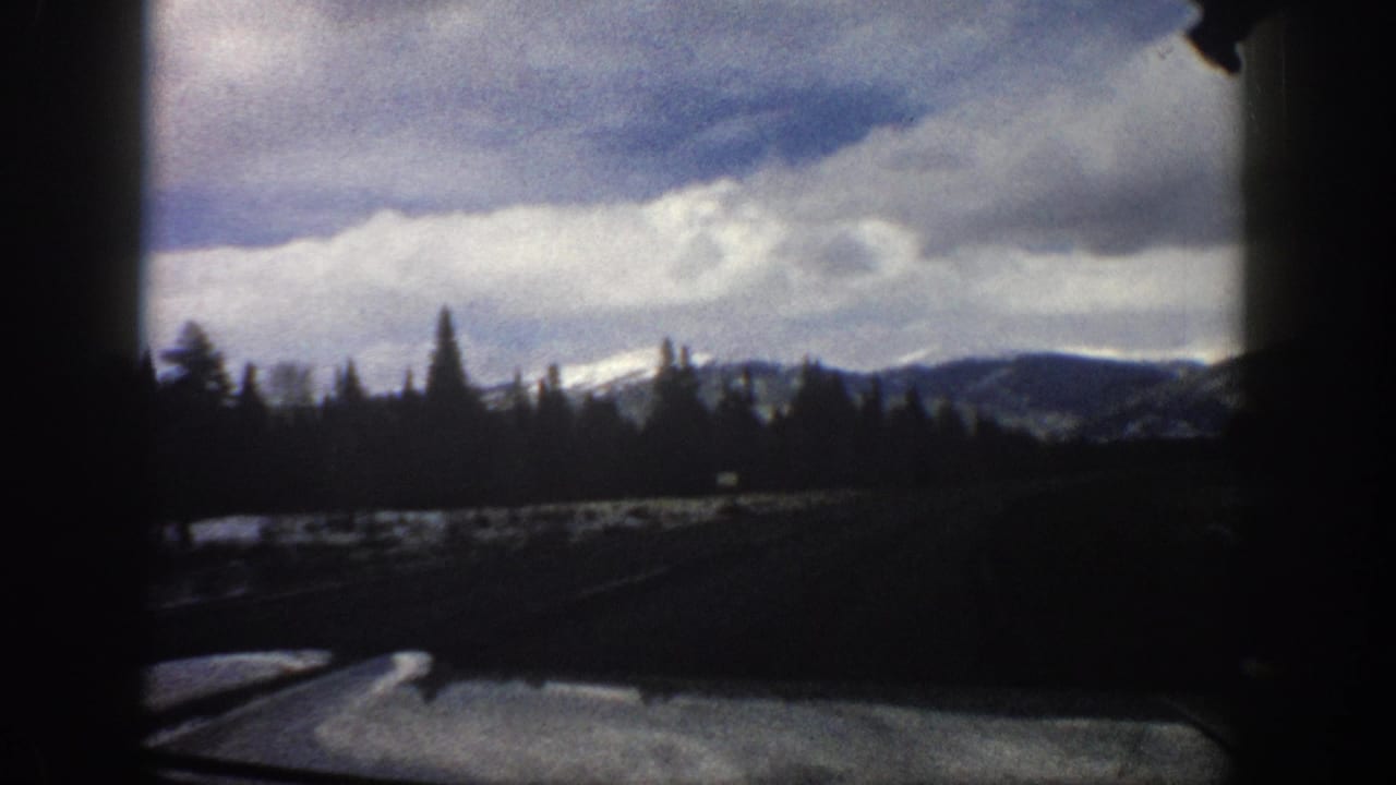 1961 Aspen-area 8mm frame showing a moving vehicle view of a snowy roadside, evergreen tree line, distant mountains, and heavy clouds.