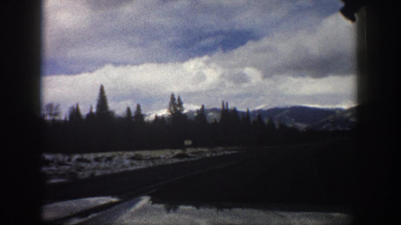 1961 Aspen, Colorado travel footage frame with a roadside snowfield, silhouetted evergreens, distant slopes, heavy clouds, and dark vehicle edges.