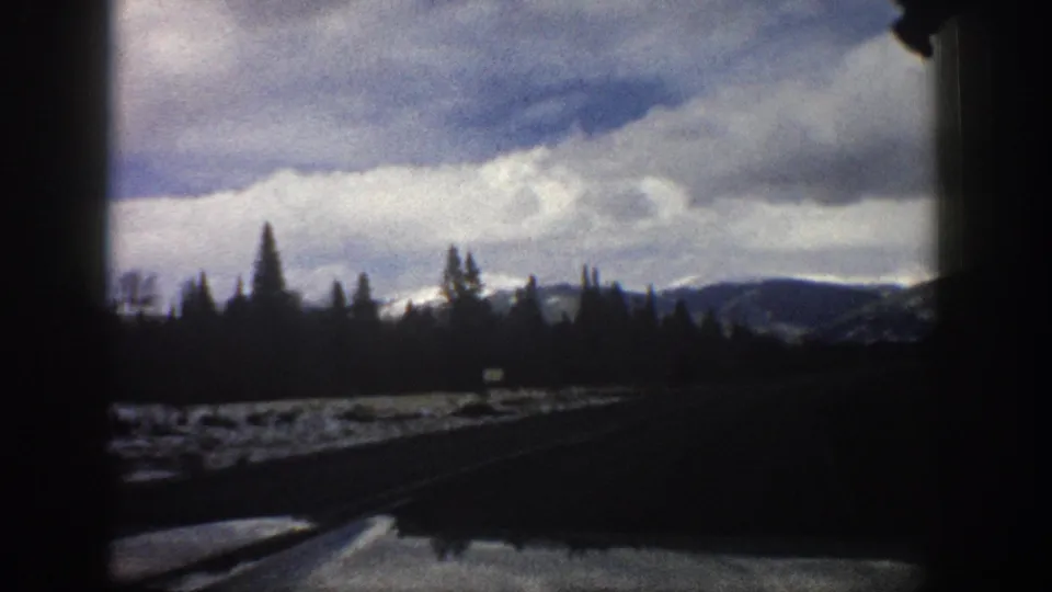 1961 Aspen, Colorado travel footage frame with a roadside snowfield, silhouetted evergreens, distant slopes, heavy clouds, and dark vehicle edges.