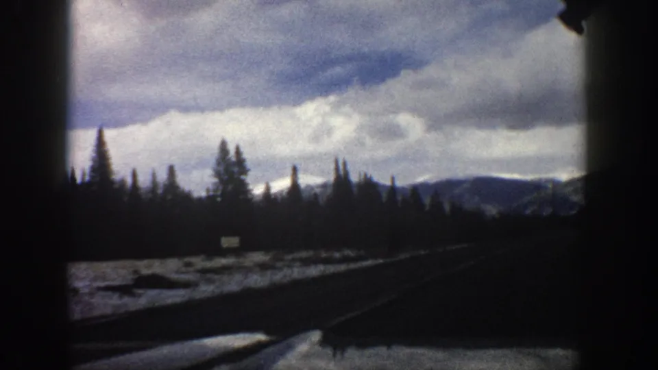A 1961 Aspen-area 8mm frame looking from a moving vehicle toward a curving road, snow patches, evergreens, and distant mountains under clouds.