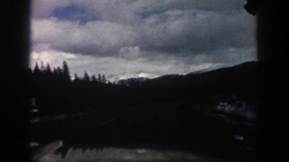 1961 Aspen, Colorado road-trip frame with a darkened moving-vehicle view of roadside snow, silhouetted evergreens, mountain slopes, and heavy clouds.