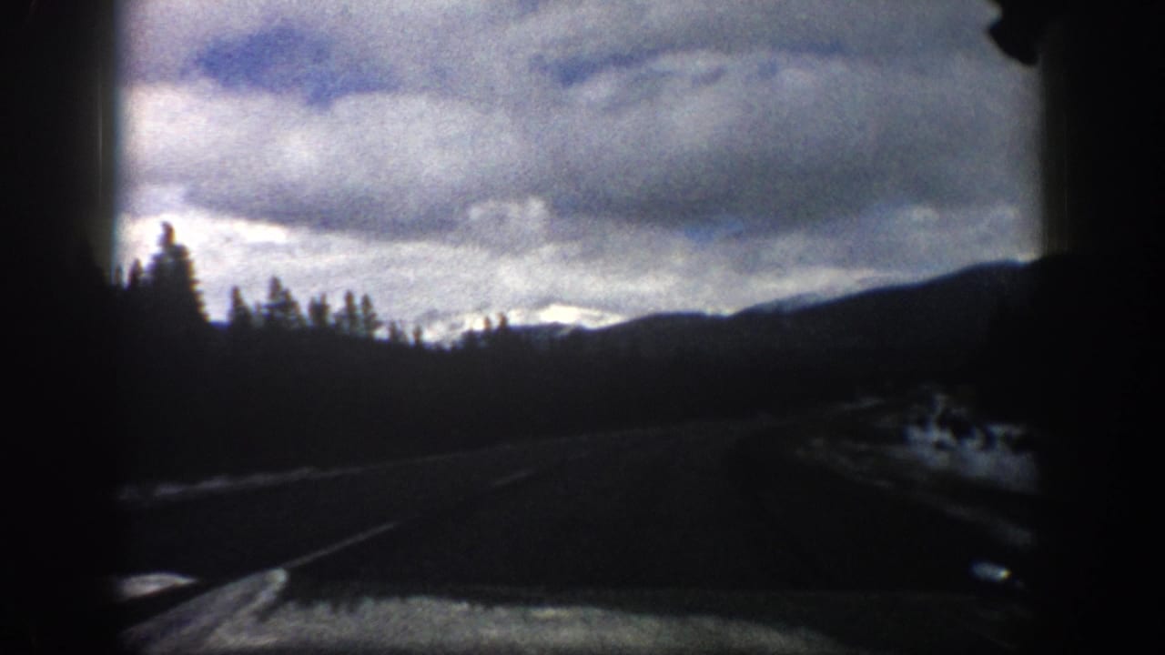 1961 Aspen-area 8mm windshield view showing a winding mountain road, evergreen silhouettes, roadside snow, and a clouded sky.