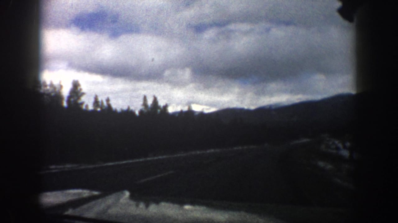 Aspen, Colorado home-movie frame with the car looking ahead toward trees, snow along the road, and low mountain ridges.