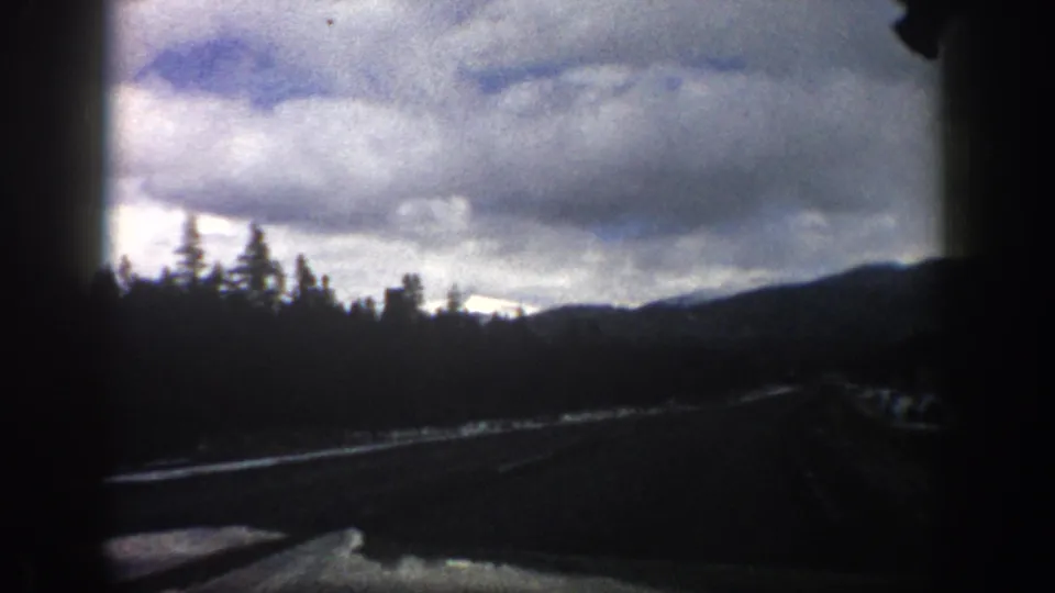 8mm Colorado road-travel frame showing the windshield-framed roadway, forested slope, distant hills, and uneven winter light.