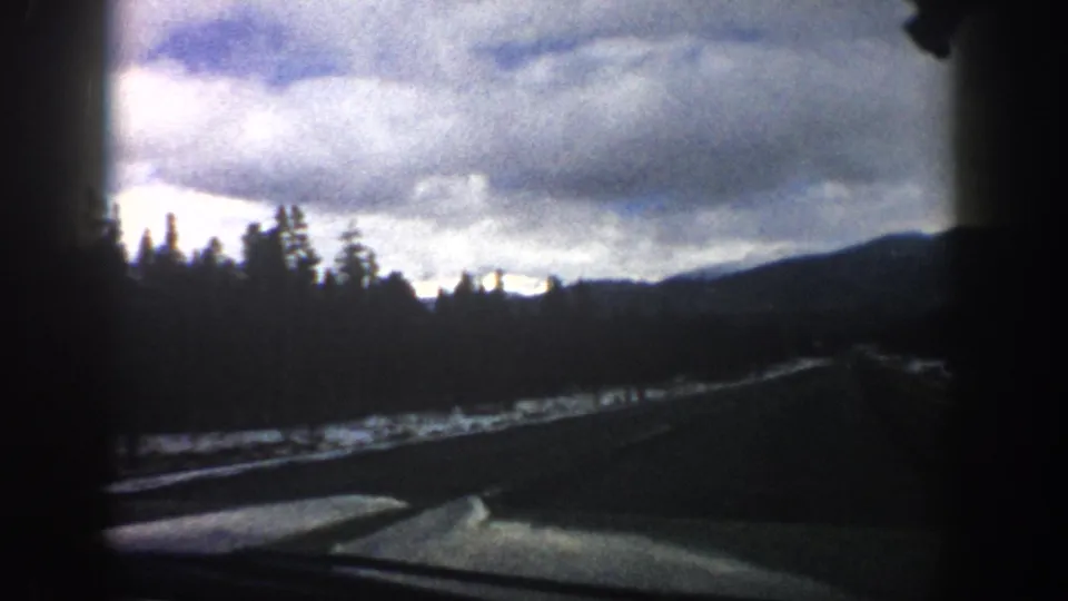 Late-frame Aspen-area windshield view with road, roadside snow, evergreen trees, and blue-gray clouds over distant mountains.