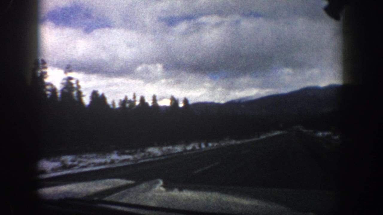 1961 Aspen-area Colorado 8mm frame showing a highway from inside a moving car, with snow patches, evergreen hills, and heavy clouds.