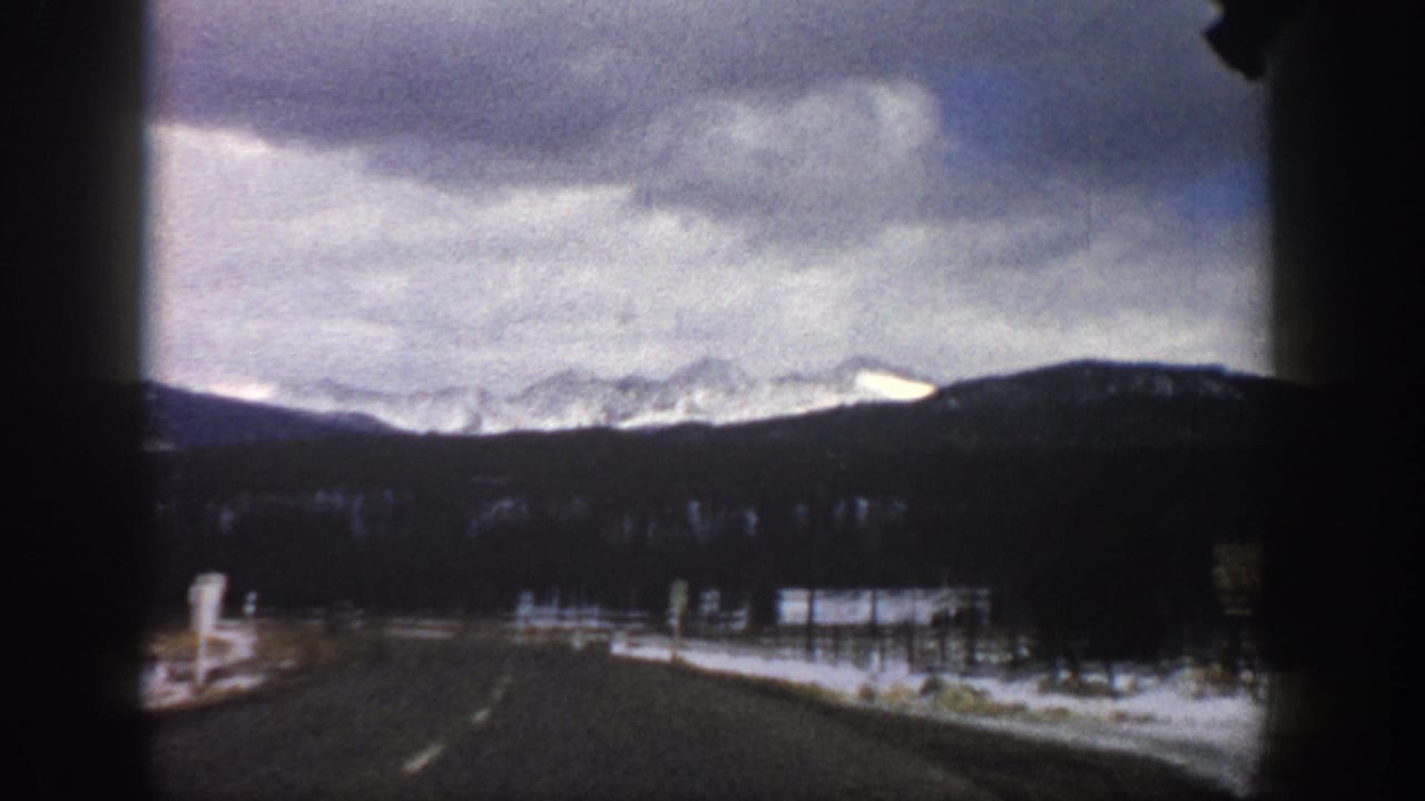 Roadway leading toward distant snowy peaks under a cloudy sky, framed by the car interior.