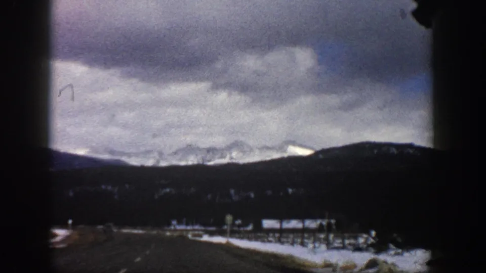 Aspen, Colorado 1961 home-movie frame of a highway stretching toward low mountains and snowy peaks, with visible 8mm grain.