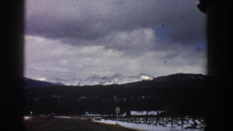 Final 1961 Aspen-area driving frame with a dark windshield edge, highway foreground, evergreen slopes, snow patches, and cloudy mountain sky.