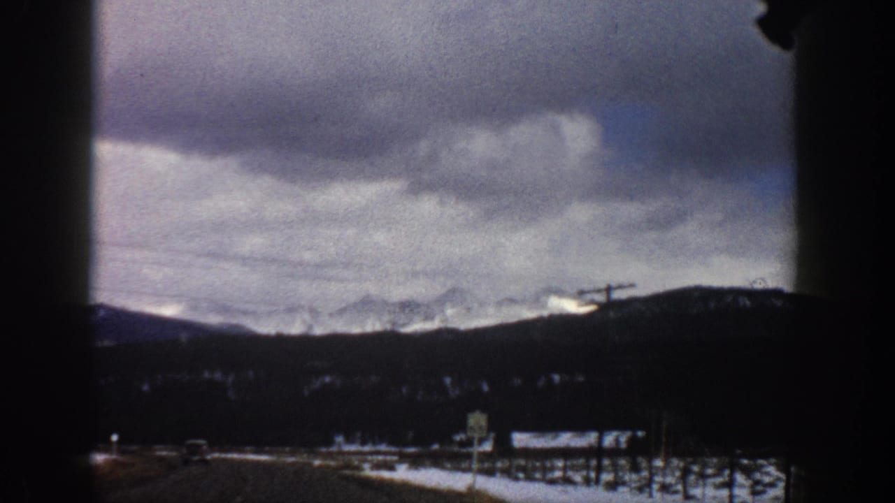 1961 Aspen-area winter road view from a moving vehicle, with snowy fields, distant mountains, utility poles, and heavy clouds.