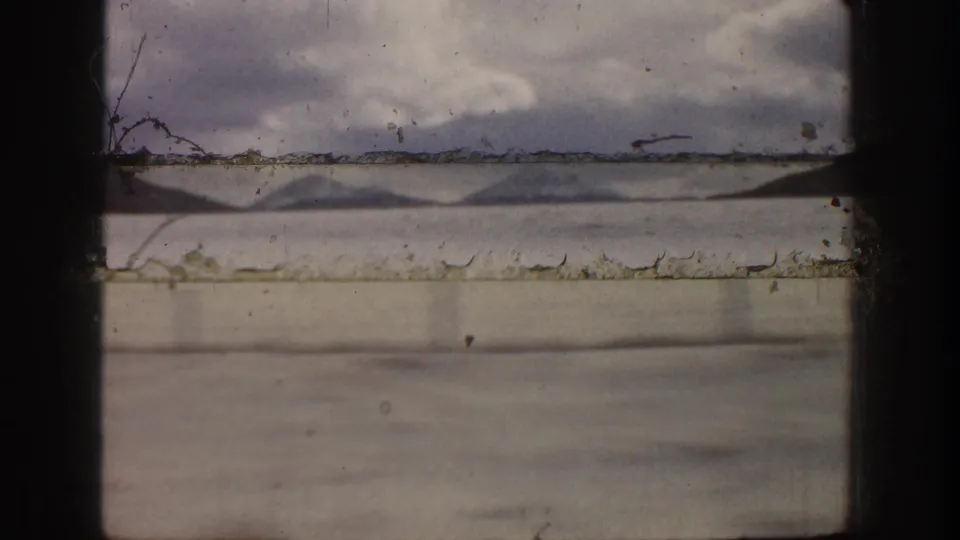 Faded 8mm home-movie frame of a mountain-and-water landscape near Aspen, with dust, scratches, and gate-edge marks visible.