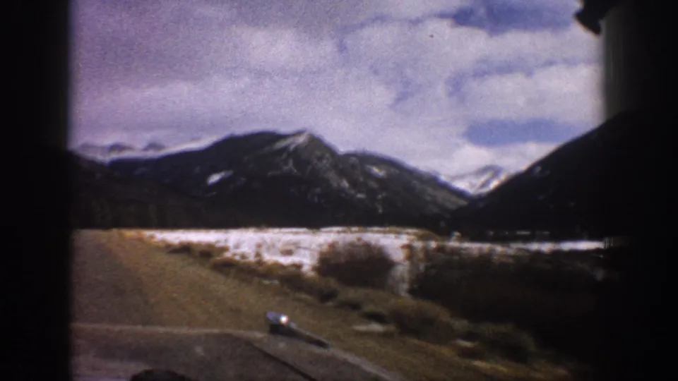 1961 Aspen-area road view from a vehicle, with snow-dusted mountains, a valley road, and cloudy sky in 8mm color.