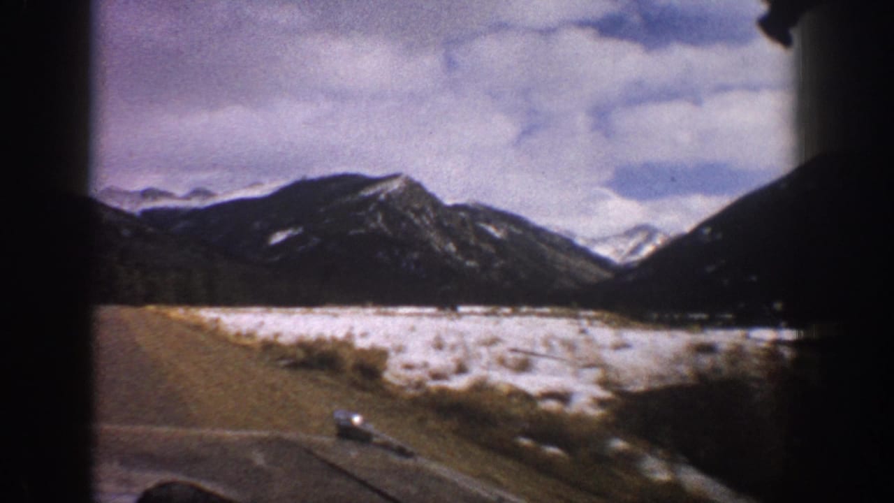 1961 Aspen-area road view from a moving car, with snowy ground or frozen water before dark mountain slopes under clouds.
