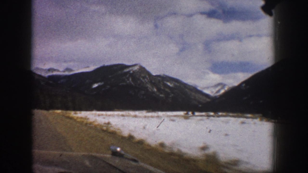 Snowy flat land and mountain slopes seen through a car window beside a roadside edge.