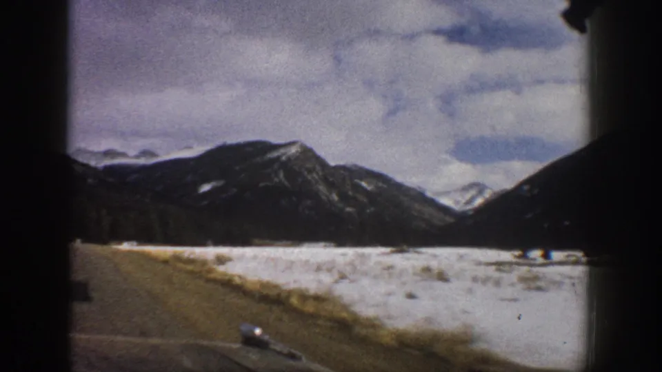 Aspen-area 8mm car-window shot from 1961, with a road shoulder, snowy open ground, and dark mountain slopes.