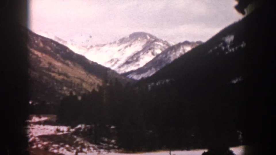 1961 Aspen, Colorado home-movie frame with snowy peaks, shadowed evergreen hillsides, and faded color film texture.