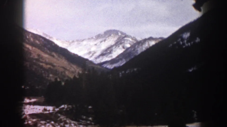 Aspen, Colorado home-movie frame with snow-bright peaks, wooded slopes, and heavy 8mm gate-edge shadows.