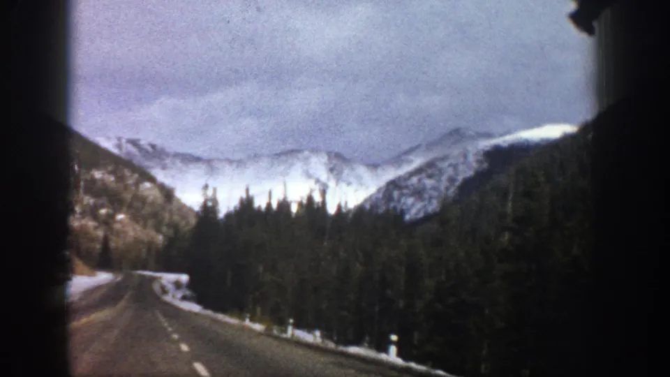 1961 Aspen-area road-travel frame showing a paved mountain road curving past snowbanks toward evergreen forest and snowy hills.