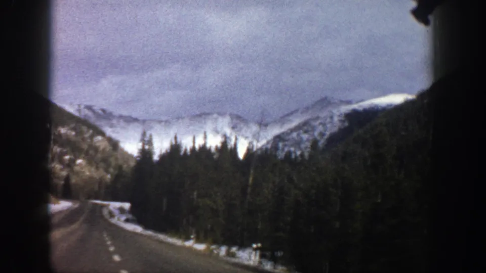 Aspen, Colorado 8mm road scene with a winding paved road, evergreen forest, snow patches, and distant snowy mountain ridges.