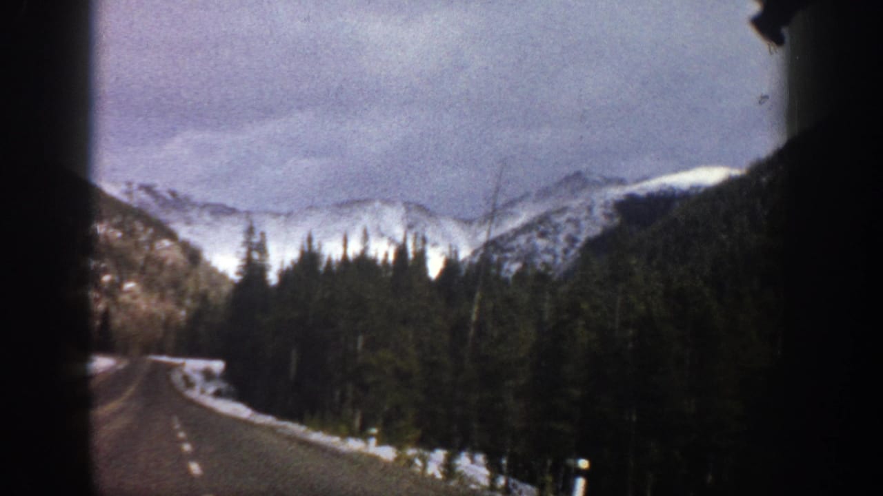1961 Aspen-area 8mm frame from inside a vehicle, showing a curving road, snowbanks, forest, and distant snow-capped mountains.