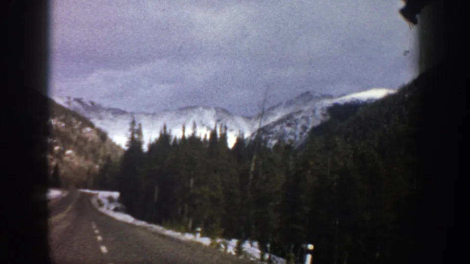 1961 Aspen-area 8mm frame from inside a vehicle, showing a curving road, snowbanks, forest, and distant snow-capped mountains.