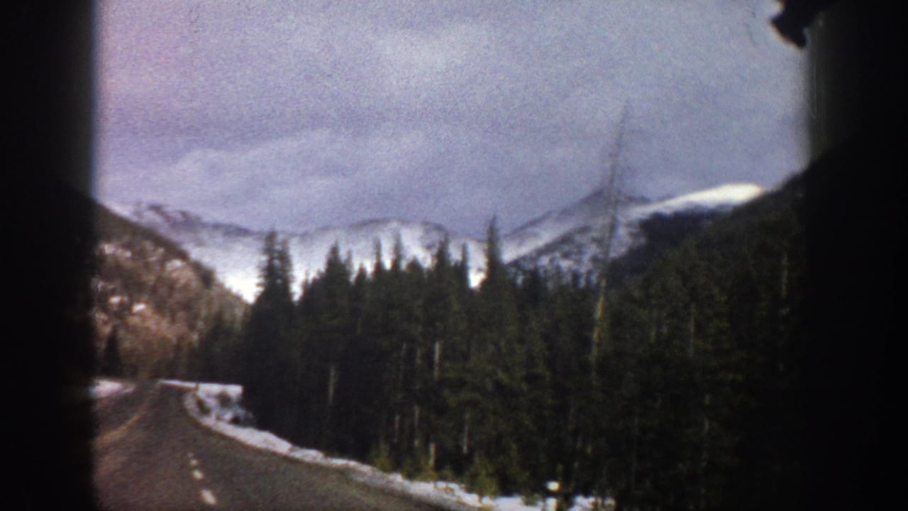 Roadside forest and snowy mountain slopes seen through dark vehicle-window edges.
