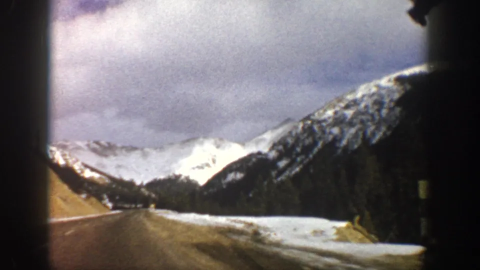 1961 8mm vehicle-window view near Aspen, showing an open paved road, snowbanks, forested slopes, and bright snowy mountains.