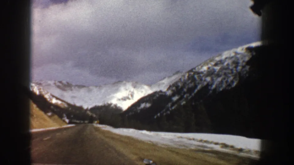 Aspen-area home-movie frame from 1961, with a straight road, snow-covered roadside, dark evergreens, and high white peaks ahead.