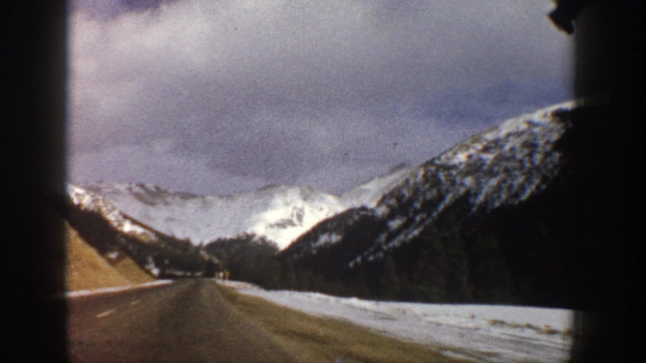 Two-lane mountain road seen through a vehicle window with snowbanks and distant peaks.