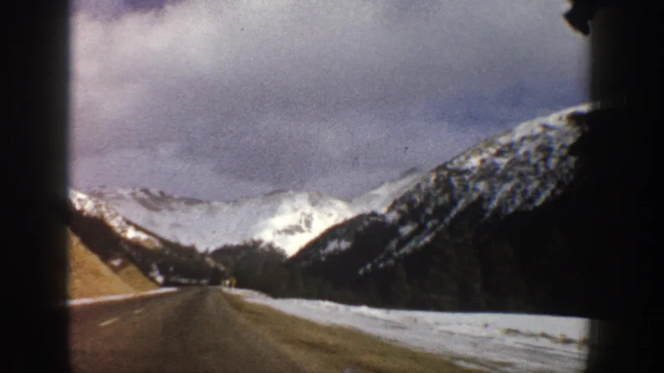 1961 Aspen-area 8mm view from inside a car, looking down a snowy mountain road toward dark slopes and white peaks.