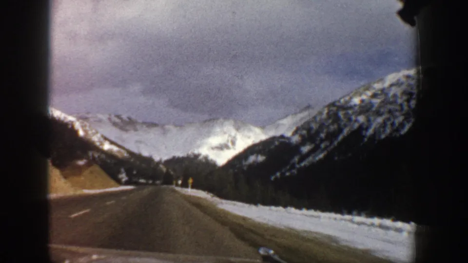 1961 8mm passenger-view frame of a snowy Colorado mountain road curving through evergreen slopes toward pale peaks.