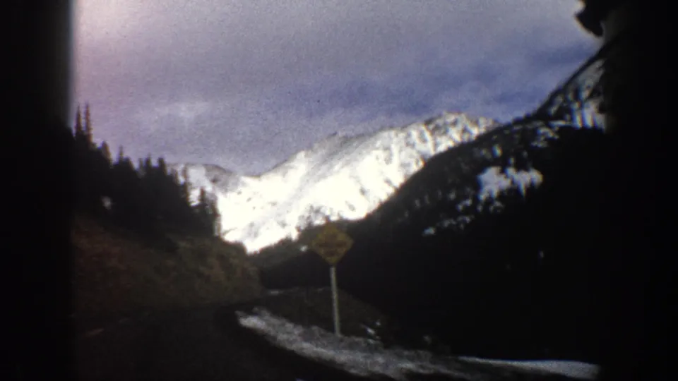 Aspen, Colorado 1961 home-movie frame showing snow-covered peaks, forested slopes, and a roadside sign from a moving car.