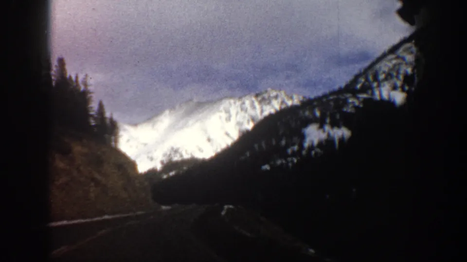 A shadowed road and roadside hillside lead toward a bright snow-covered mountain slope in 1961 Aspen home-movie footage.
