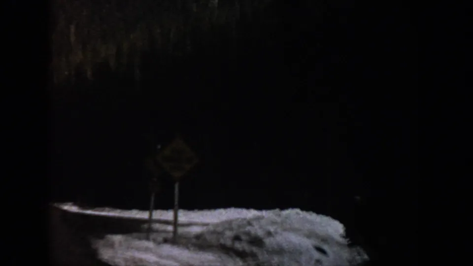 1961 Aspen-area 8mm frame showing a yellow roadside warning sign beside snowbanks and dark forest.