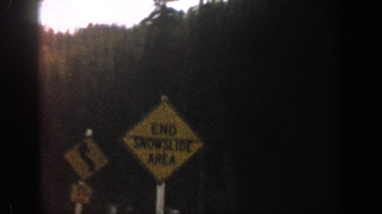 1961 Aspen-area frame with a yellow winding-road warning sign, snow at roadside, and forest beyond.