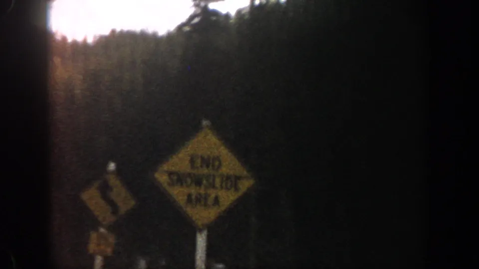 1961 Aspen-area frame with a yellow winding-road warning sign, snow at roadside, and forest beyond.